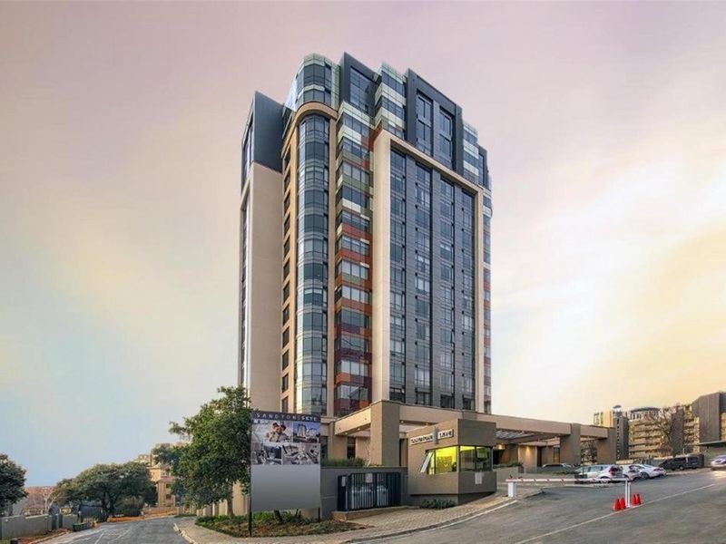 A modern high-rise apartment building featuring a glass and concrete facade, secure gated entrance, and professional landscaping.