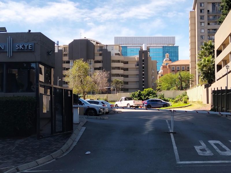 View of the Sandton Skye apartment complex entrance, featuring a security gatehouse, paved driveway, and surrounding multi-story residential buildings.