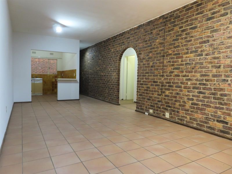 An empty open-plan living area featuring tiled floors, a prominent exposed brick wall with an arched doorway, and a pass-through window to the kitchen.