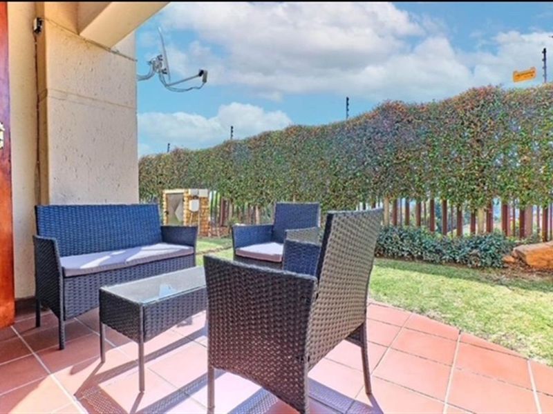 A covered patio area featuring tiled flooring, overlooking a private garden space with established hedging and perimeter security fencing.