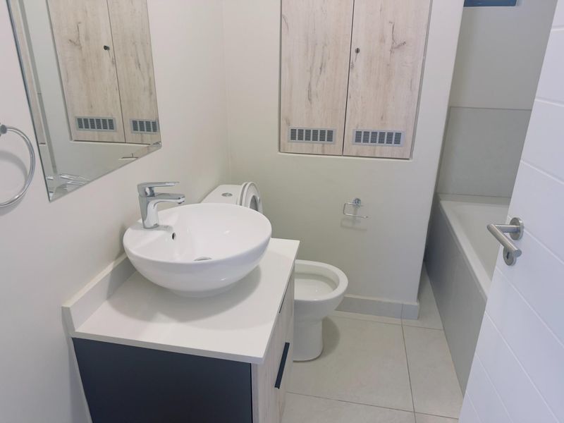 A modern, clean bathroom featuring a vessel sink vanity, toilet, and bathtub with neutral tiling and built-in storage cabinetry.