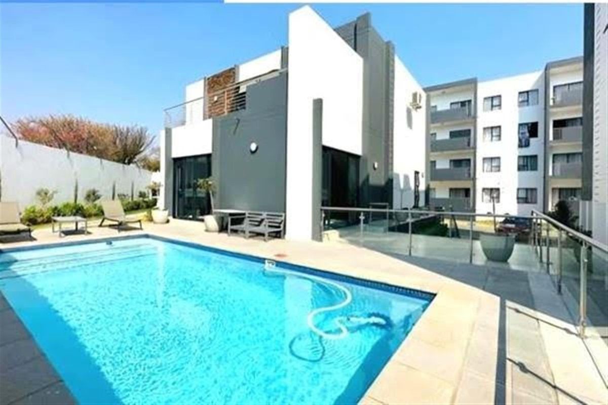 A modern apartment complex pool area featuring a clear swimming pool, paved decking, and contemporary architectural facade with glass balustrades.