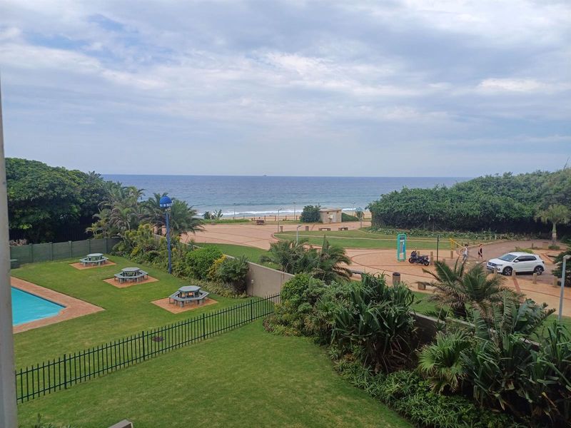 Elevated view overlooking a communal garden, swimming pool area, and the nearby coastline with direct beach access.