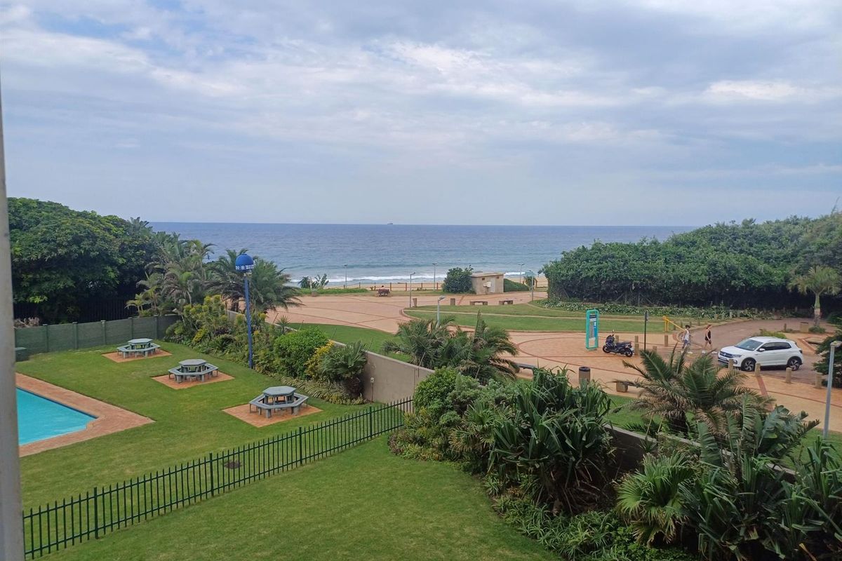 Elevated view overlooking a communal garden, swimming pool area, and the nearby coastline with direct beach access.