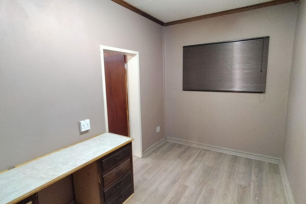 A vacant bedroom featuring light-toned laminate flooring, a built-in desk unit, and venetian blinds, offering a neutral space ready for personalization.