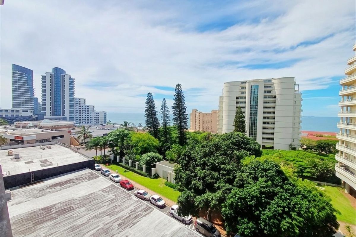 Elevated urban view overlooking surrounding apartment buildings, mature trees, and distant coastal scenery.