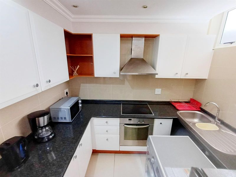 A compact kitchen featuring white cabinetry, dark granite countertops, a built-in oven, electric hob, stainless steel extractor fan, and a stainless steel sink.