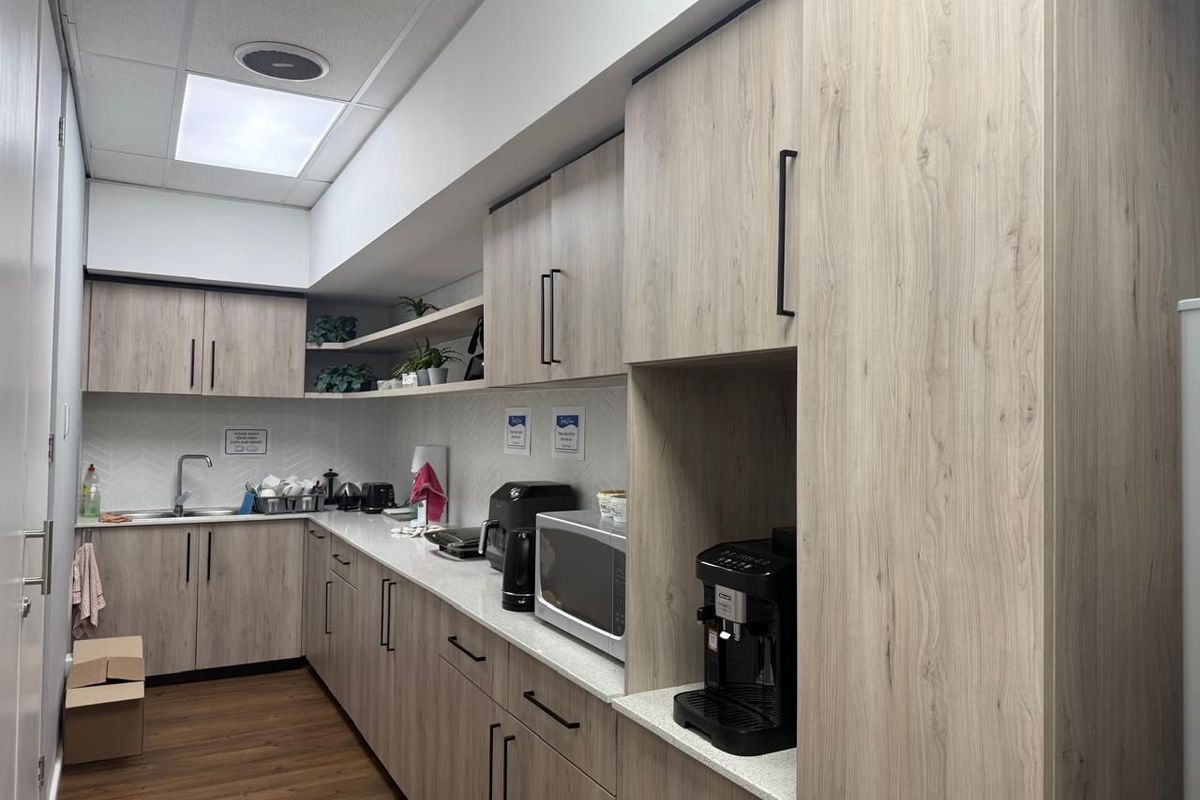 A modern commercial kitchenette featuring light wood-grain cabinetry, stone-look countertops, a stainless steel sink, and integrated shelving in a clean, functional workspace.