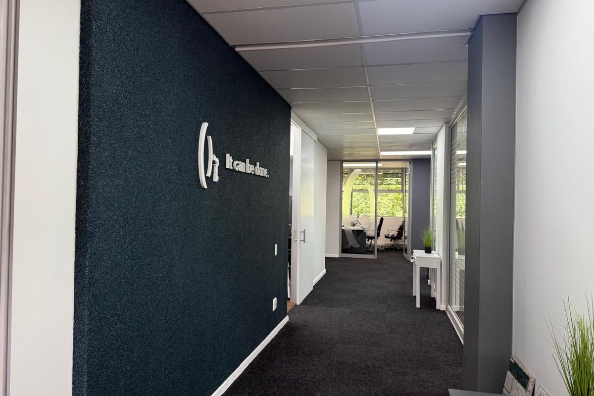 A modern commercial office hallway featuring dark carpeting, a textured feature wall, and glass-partitioned office spaces with natural light.