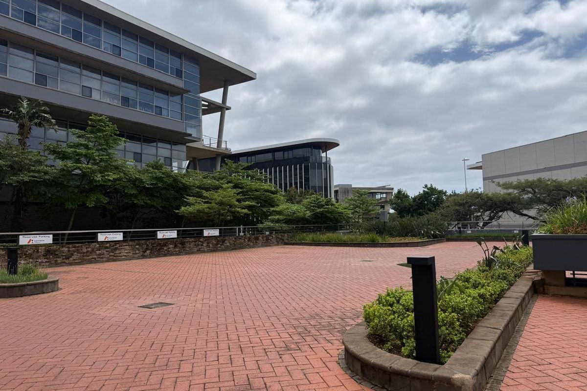 A paved outdoor courtyard area featuring landscaped garden beds and modern commercial office buildings in the background.