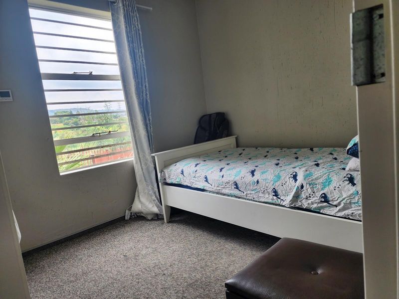 A bedroom featuring carpeted flooring and a louvered window with natural light, showing signs of wear on the wall surfaces.