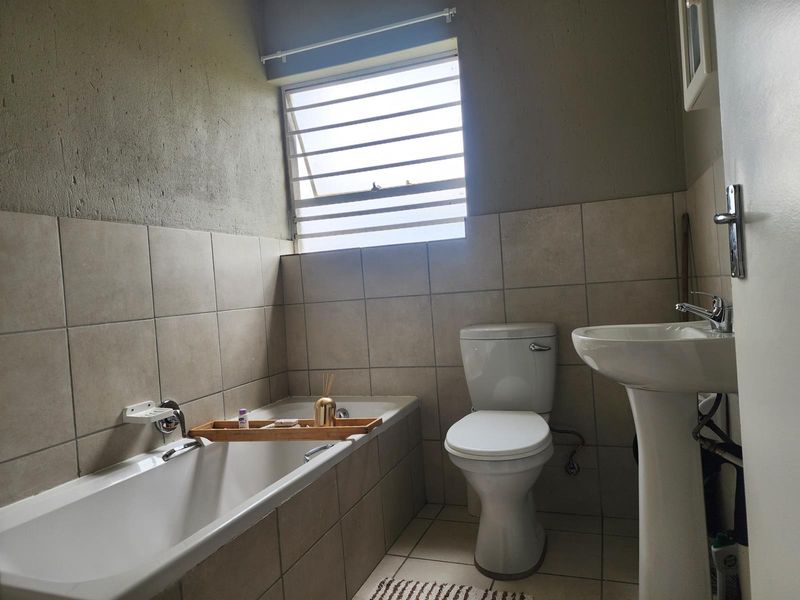 A functional bathroom featuring a bathtub, pedestal basin, and toilet with neutral wall and floor tiling and a louver window for ventilation.