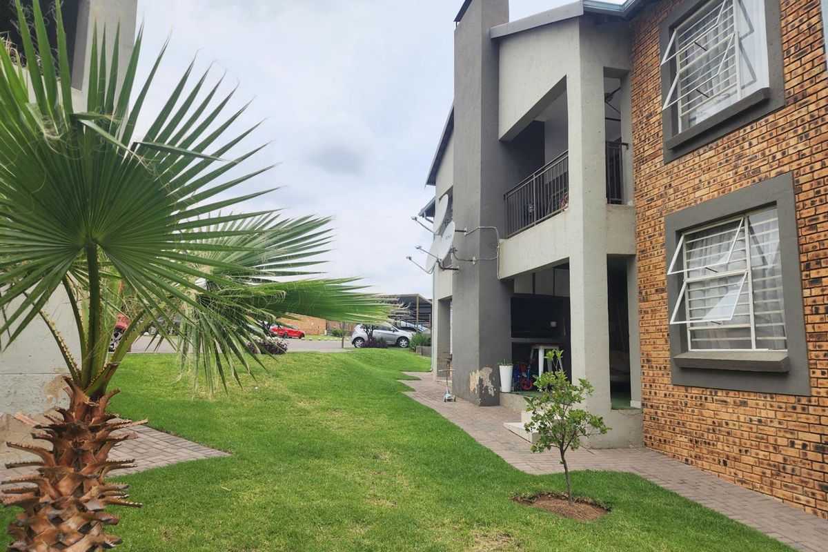 Exterior view of the apartment building featuring a brick facade, a ground-floor patio with a built-in braai, and a shared garden area.