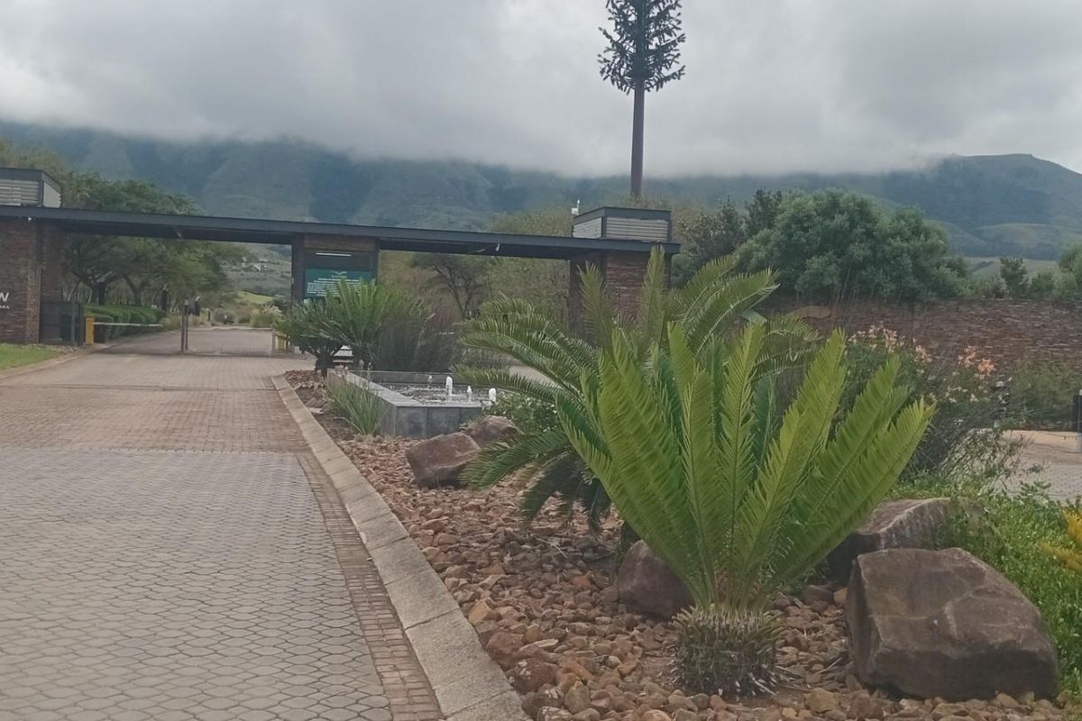 The entrance to the residential estate features a secure gatehouse, paved driveway, and landscaped gardens set against a scenic mountain backdrop.