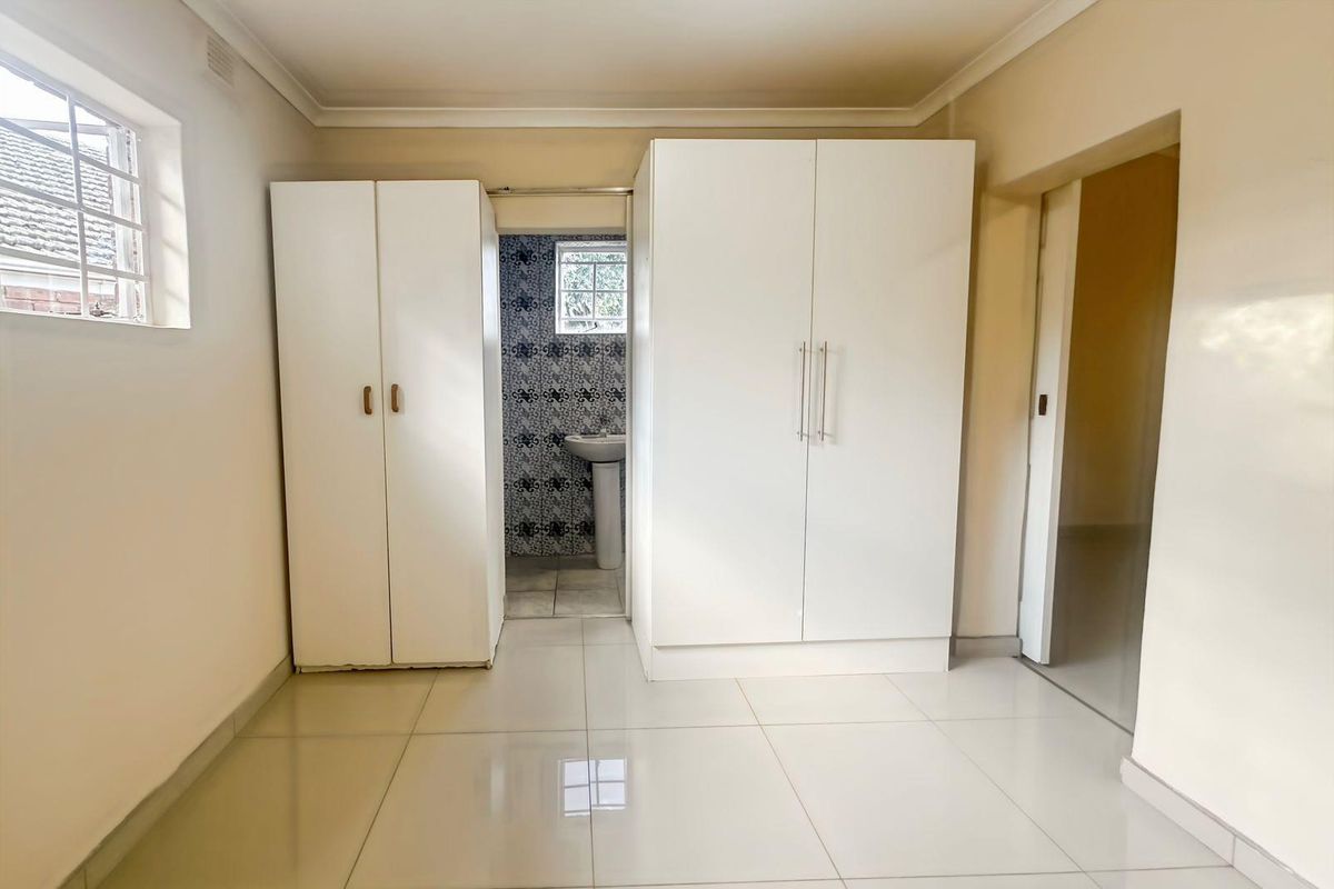 A bright, empty bedroom featuring tiled flooring, built-in cupboards, and an adjoining en-suite bathroom area with a pedestal basin.