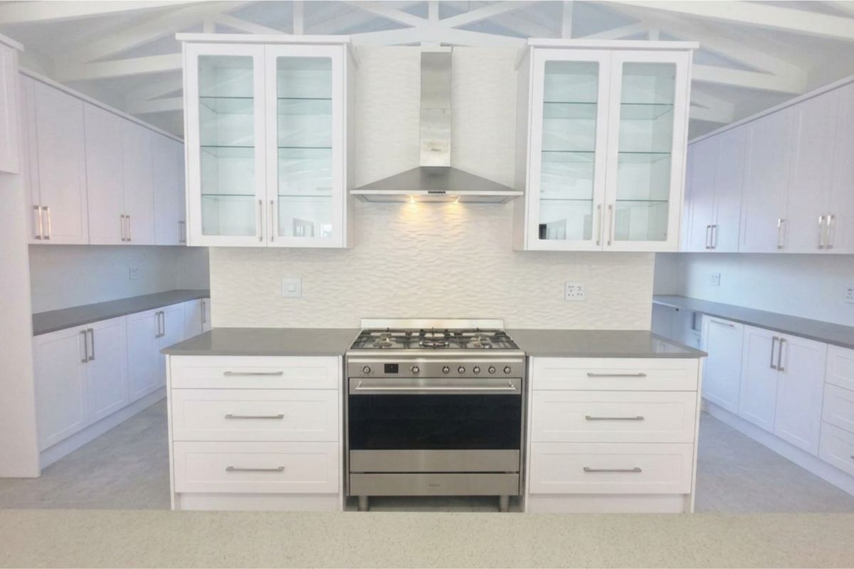 A modern, bright kitchen featuring white cabinetry, stone countertops, a stainless steel gas stove with extractor fan, and a textured tiled backsplash.
