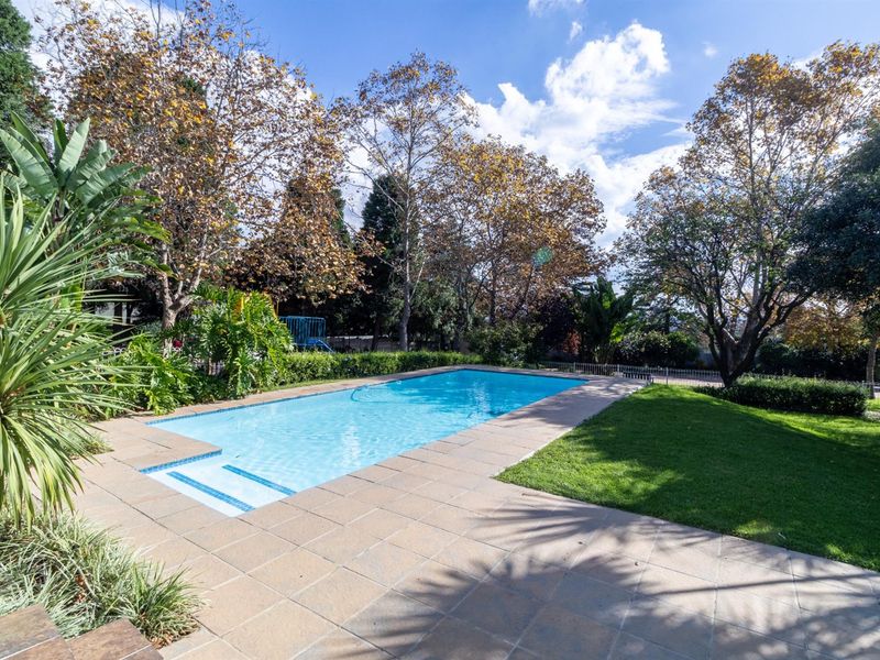A well-maintained outdoor swimming pool area featuring a tiled surround, manicured lawn, and established garden landscaping.