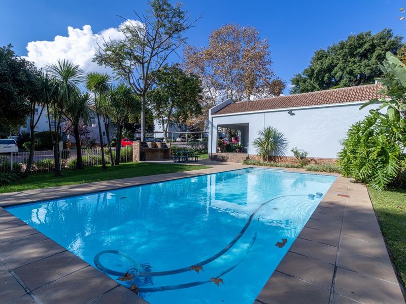 A well-maintained outdoor pool area featuring a tiled surround, built-in brick braai, and a covered patio space within a landscaped garden setting.