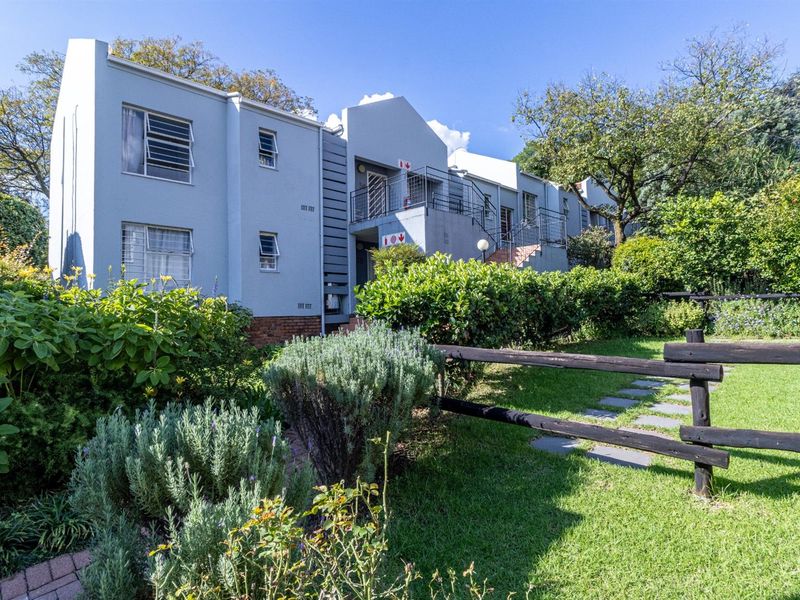 A well-maintained apartment building exterior featuring landscaped gardens, paved pathways, and secure metal railings, set within a lush green residential environment.