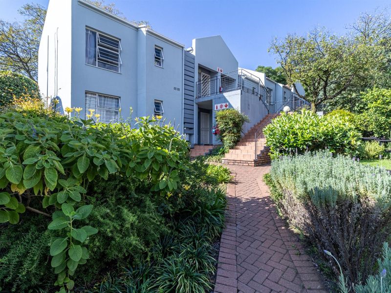 Front exterior view of a multi-story apartment building featuring a brick paved walkway, landscaped garden, and external staircase access.
