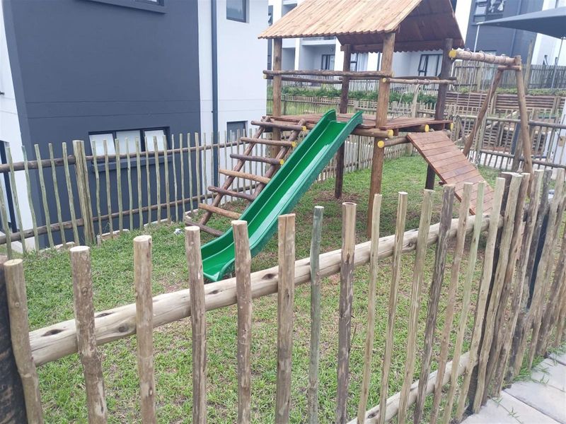 A communal garden area featuring a wooden children's play structure with a slide, enclosed by rustic wooden pole fencing.
