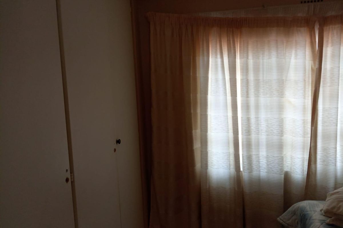 A bedroom featuring built-in cupboards and a window with curtains, showing signs of age and wear.