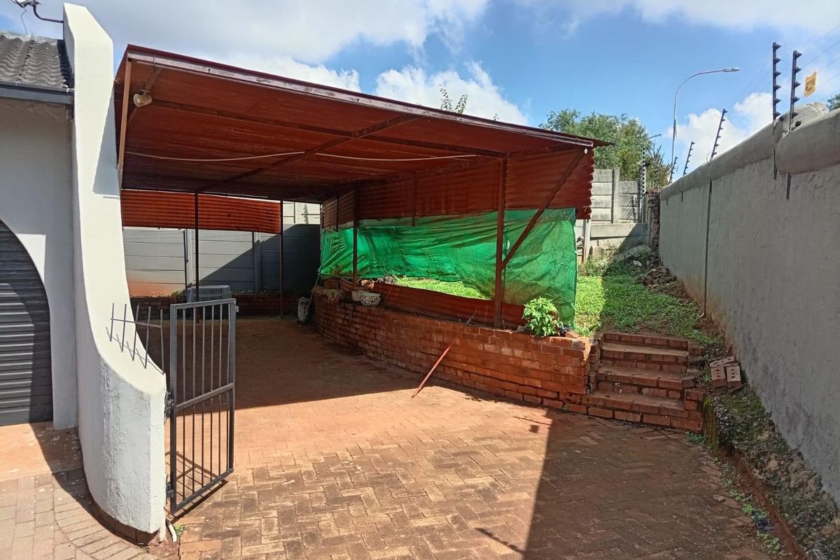 An outdoor area featuring a paved driveway, a metal-roofed carport structure, brick retaining walls, and perimeter security fencing.