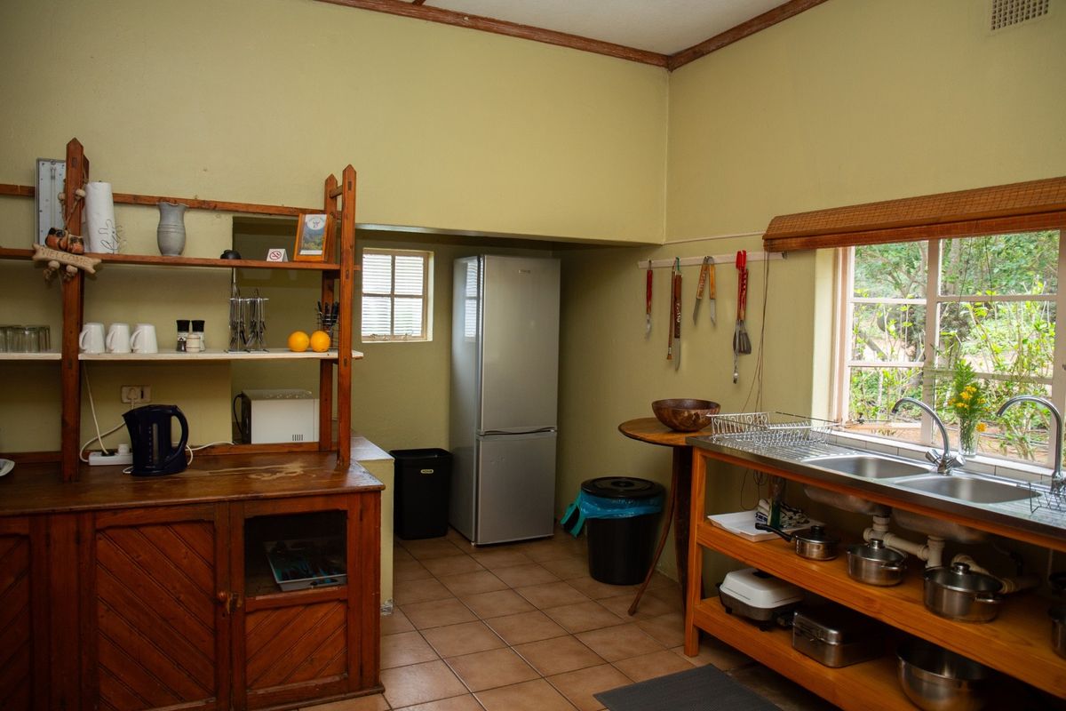 A functional kitchen area featuring a stainless steel double sink, open wooden shelving, and tiled flooring with a view of the garden.