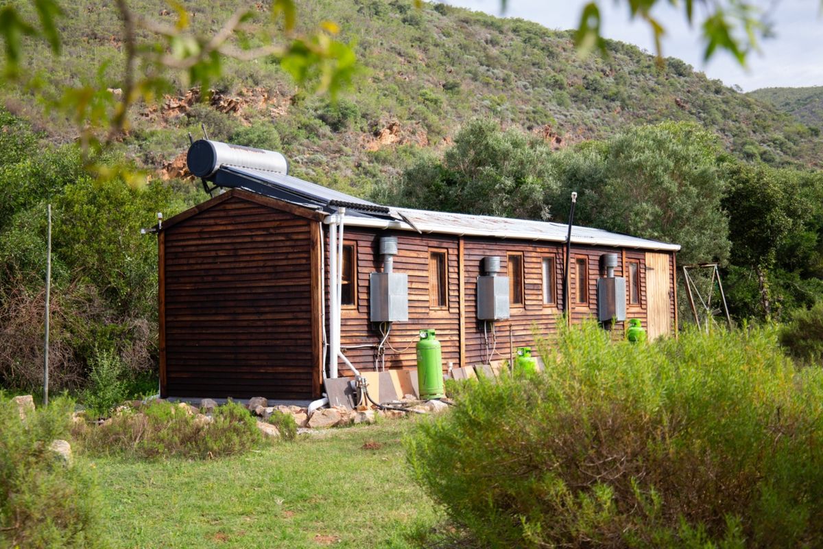 A rustic timber-clad cabin set in a natural landscape, featuring a solar water heating system and external gas geyser installations.