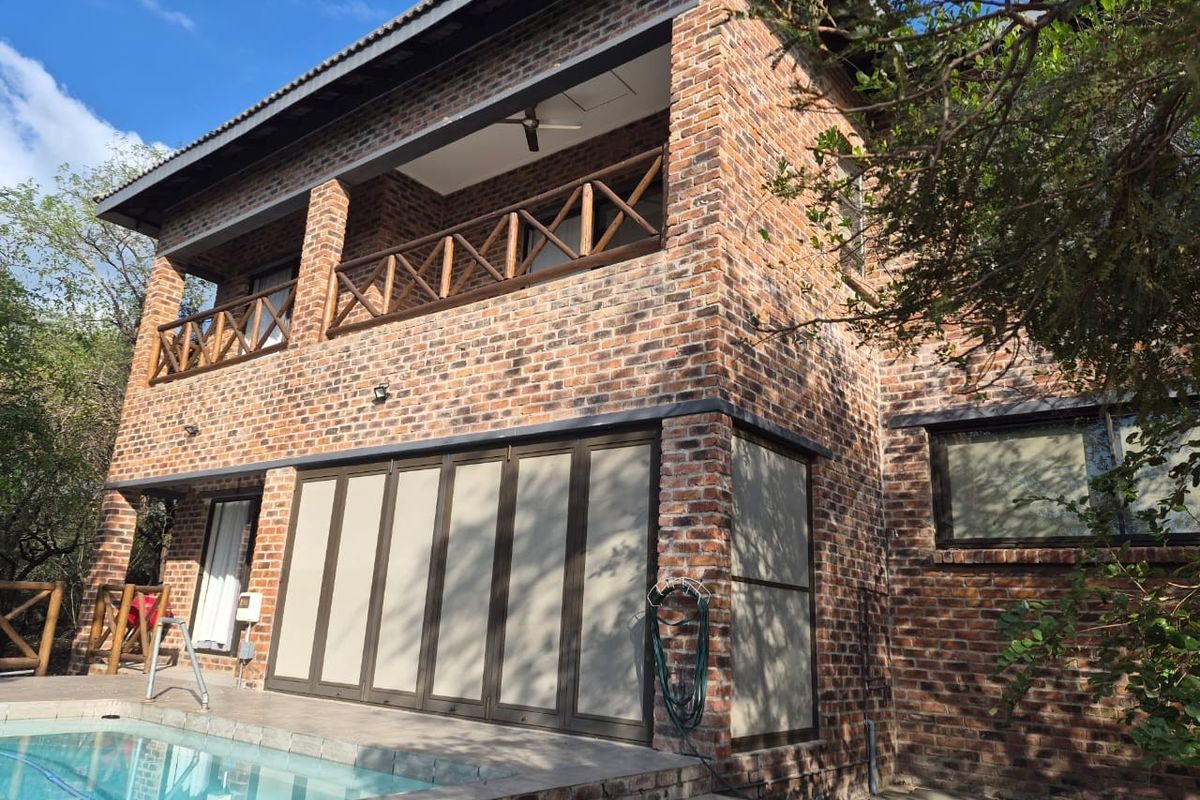 Two-story face-brick house featuring a covered upper balcony with wooden railings and a ground-level patio area adjacent to a swimming pool.