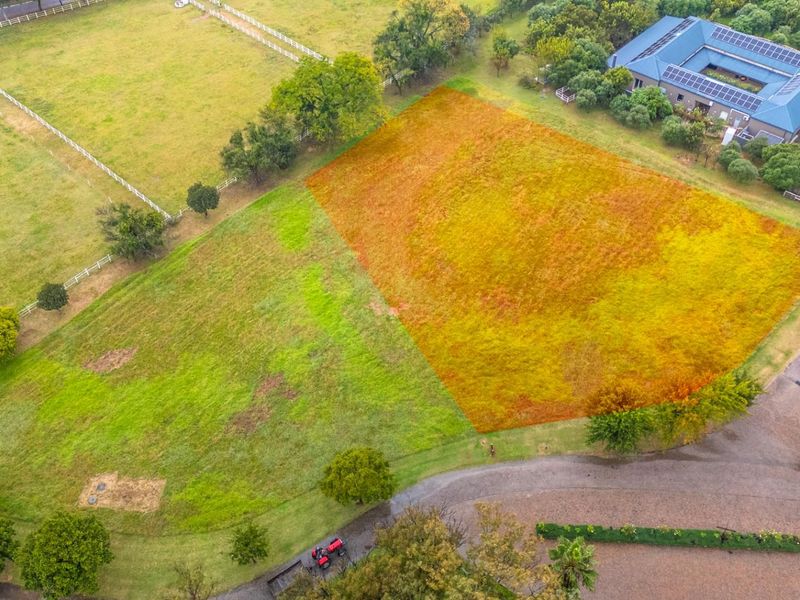 An aerial view of a vacant residential plot featuring established grass, mature trees, and clear boundary lines within a developed estate setting.