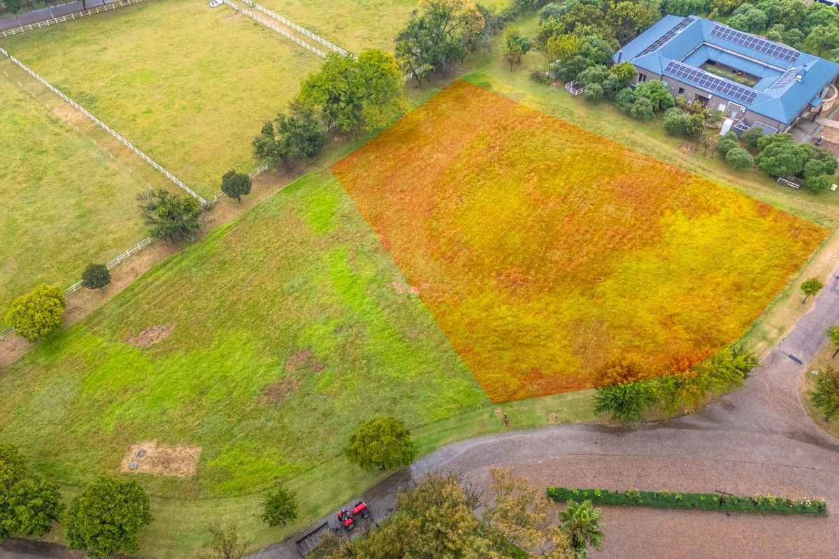 An aerial view of a vacant residential plot featuring established grass, mature trees, and clear boundary lines within a developed estate setting.