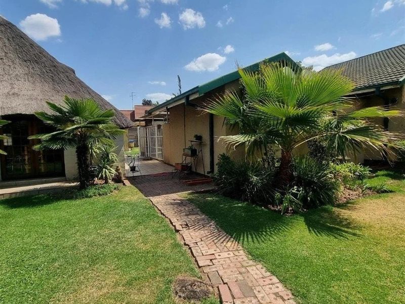 A well-maintained rear garden featuring a brick pathway, lush lawn, and a combination of thatched and tiled roof structures.