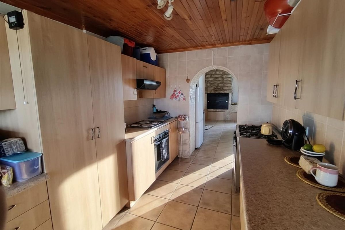 A functional kitchen featuring light wood-finish cabinetry, tiled countertops, a built-in oven and hob, and a timber-clad ceiling, leading to an arched indoor braai area.