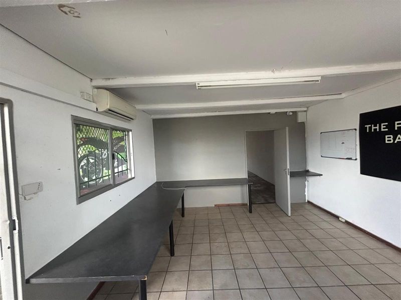 A functional commercial office space featuring tiled flooring, built-in desk surfaces, a wall-mounted air conditioning unit, and a security-screened window.