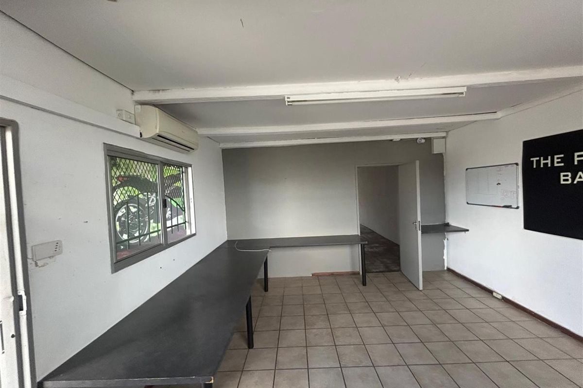 A functional commercial office space featuring tiled flooring, built-in desk surfaces, a wall-mounted air conditioning unit, and a security-screened window.