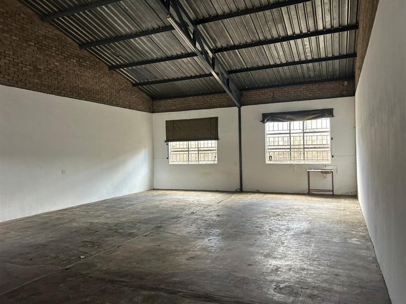 An open-plan commercial space featuring high exposed steel truss ceilings, concrete flooring, and large windows with security bars.