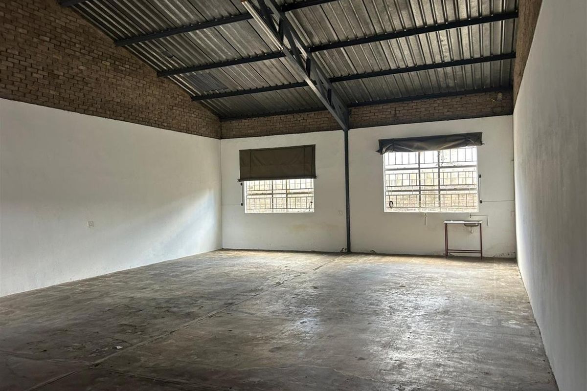 An open-plan commercial space featuring high exposed steel truss ceilings, concrete flooring, and large windows with security bars.