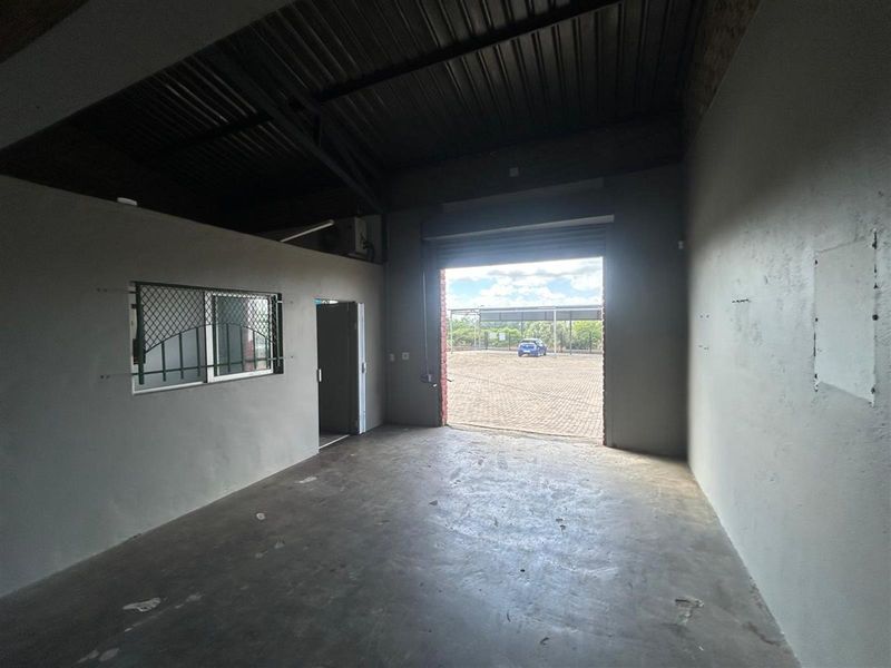 An open-plan commercial utility space featuring exposed industrial ceiling, concrete flooring, a security-grilled window, and a large roller shutter door access.