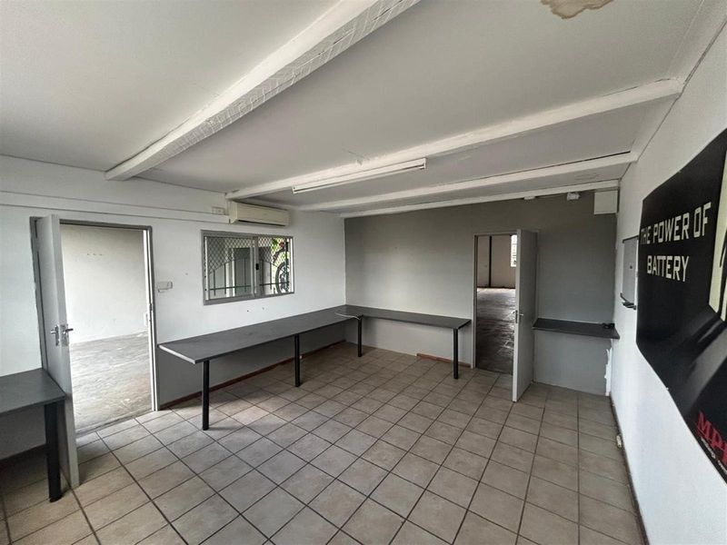A functional commercial office space featuring tiled flooring, built-in desk surfaces, a wall-mounted air conditioning unit, and direct access to adjacent rooms.