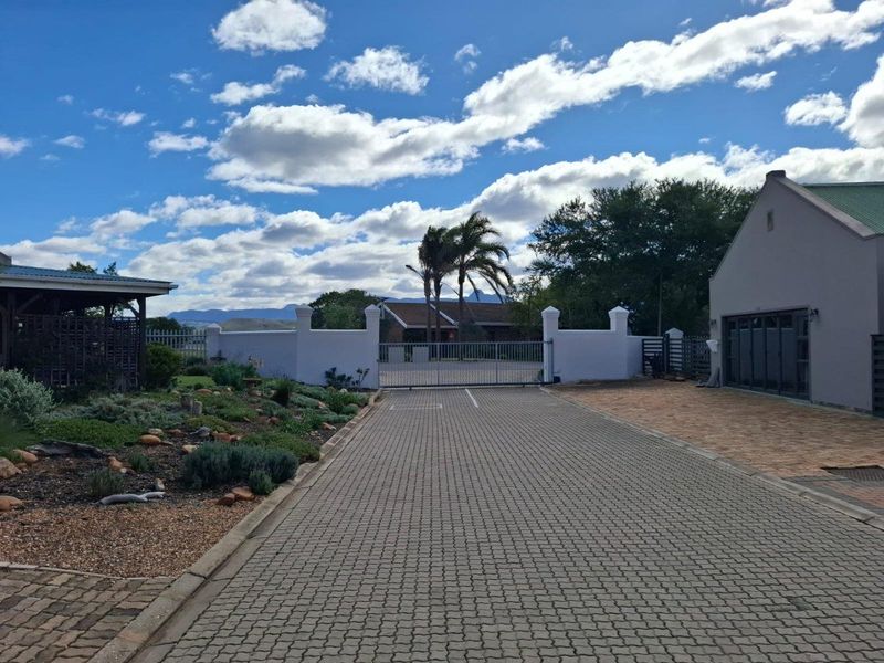 A well-maintained residential entrance featuring a paved driveway, secure perimeter wall with a metal gate, and landscaped garden area.