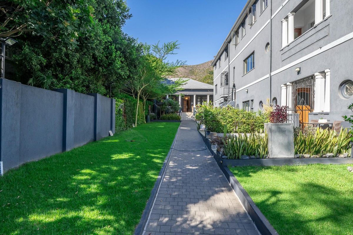A well-maintained exterior walkway leading through a landscaped garden area alongside the apartment building, featuring paved paths and established greenery.