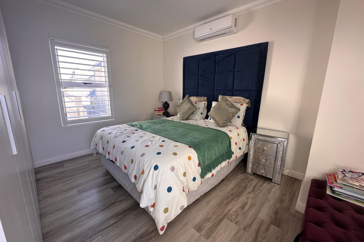 A well-maintained bedroom featuring modern wood-look flooring, a wall-mounted air conditioning unit, and a large window with fitted shutters.