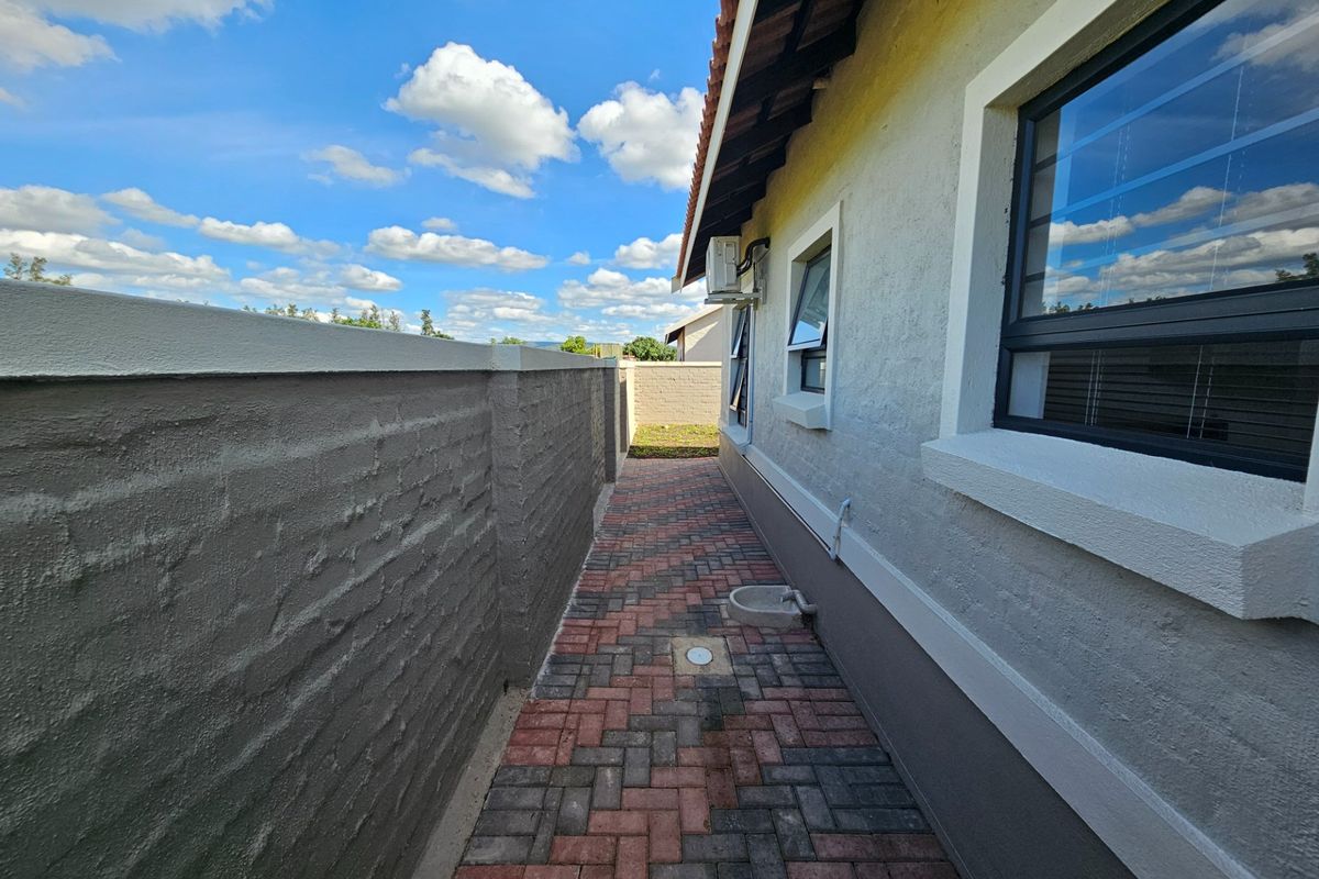 A side exterior walkway featuring paved brick flooring, a boundary wall, and an external air conditioning unit mounted on the house wall.