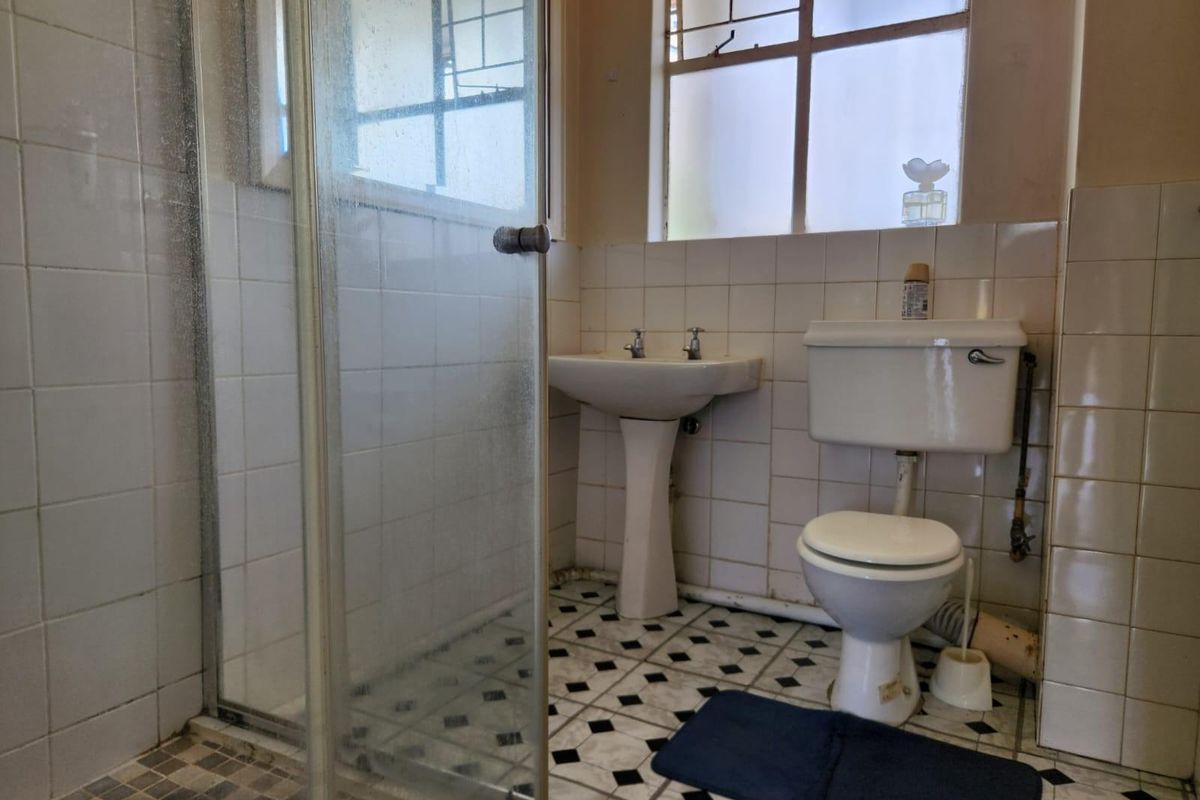 A dated bathroom featuring a pedestal basin, toilet, and a glass-enclosed shower with original wall and floor tiling.