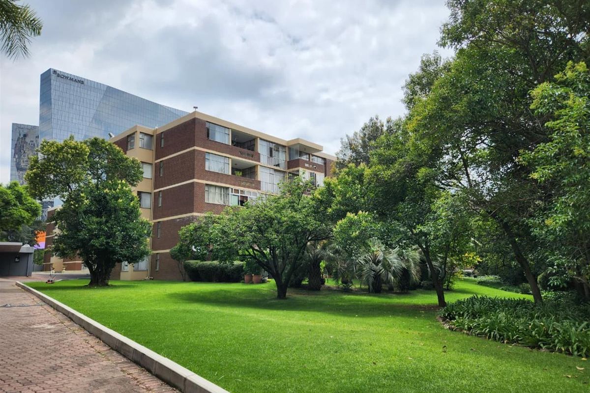 A view of a multi-story brick apartment building set within a well-maintained garden with mature trees and manicured lawns.