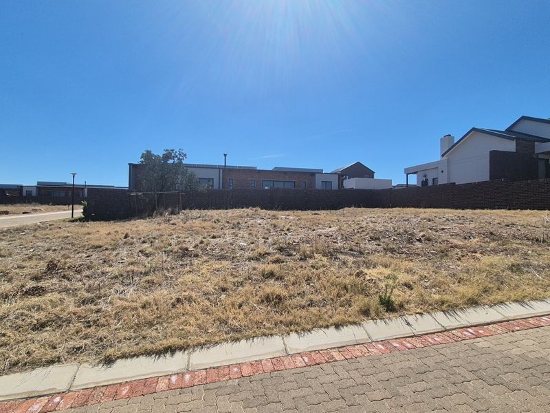 A vacant residential plot featuring dry grass, a brick boundary wall, and paved street curbing in a developed estate setting.