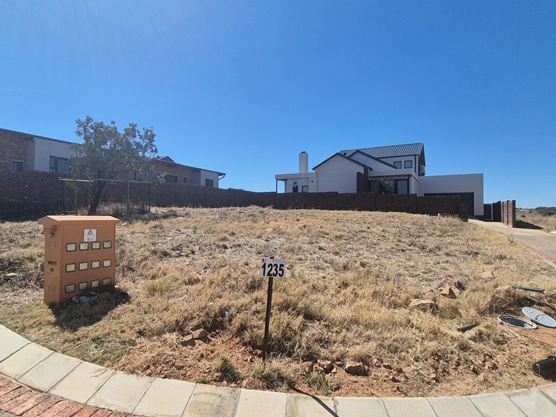 A vacant residential plot featuring a gentle slope, an electrical utility box, and established brick boundary walls in a developed estate setting.