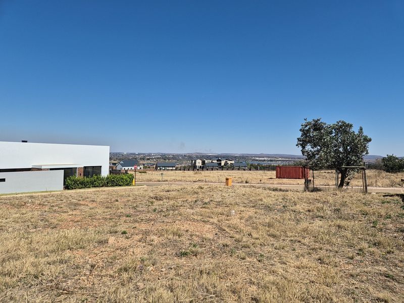 A view of a vacant residential plot featuring dry grass, perimeter fencing, and surrounding neighborhood development.