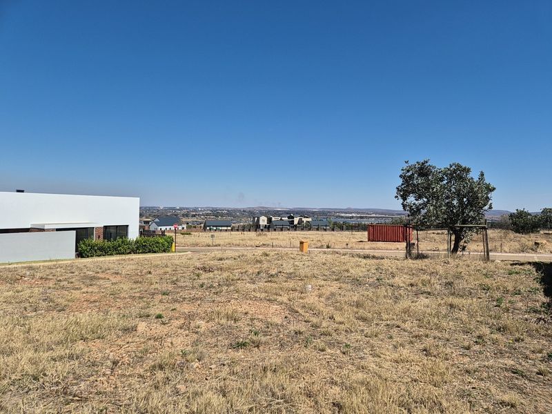 A view of a vacant residential plot featuring dry grass, boundary fencing, and surrounding neighborhood development under a clear blue sky.